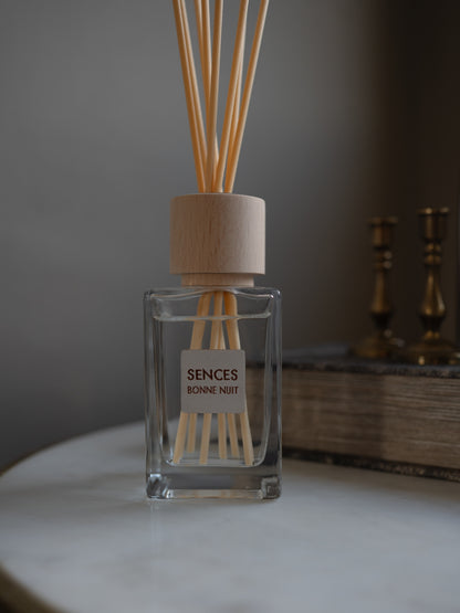 A clear glass reed diffuser bottle with a light wood cap and natural reeds, labeled &