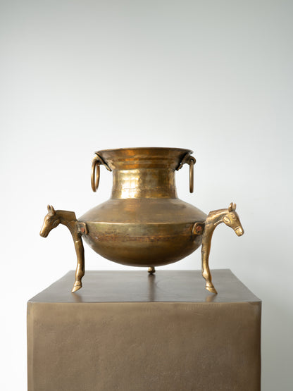 An antique-style bronze metal cauldron or pot with horse-shaped side handles, displayed on a square pedestal against a light gray background.