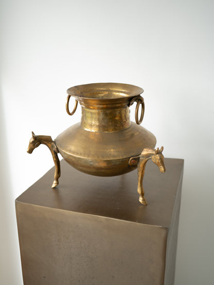 An antique-style brass pot with horse-shaped legs and ring handles, displayed on a dark metal pedestal against a plain light gray background.