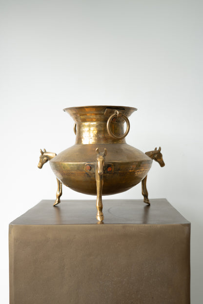 An antique-style brass or bronze trika pot with horse-head handles and ring handles, displayed on a square pedestal against a light gray background.