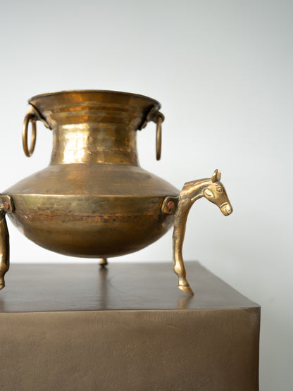 A decorative bronze-toned metal vessel with a rounded body, ring handles on the sides, and a horse-shaped leg support on the right, displayed on a metallic pedestal against a plain light background.