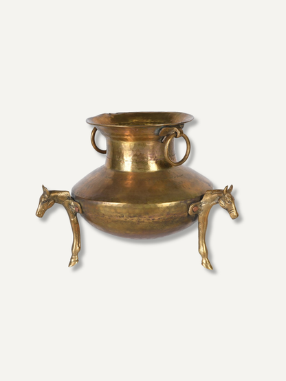 An antique-style brass pot with a wide flared rim, two circular ring handles, and three horse-shaped legs, shown against a plain off-white background.