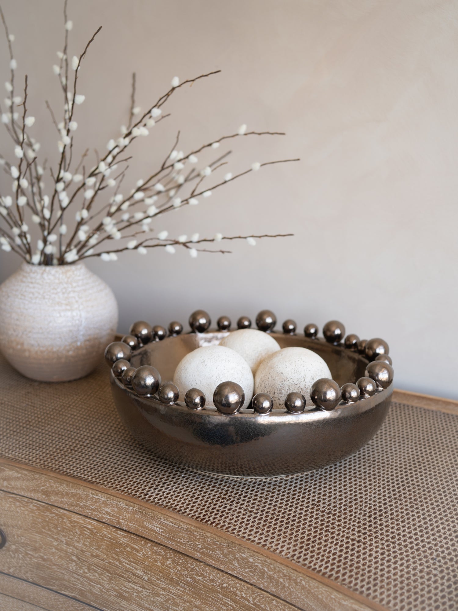 Decorative bowl with spherical balls on a wooden surface with a vase of branches in the background.