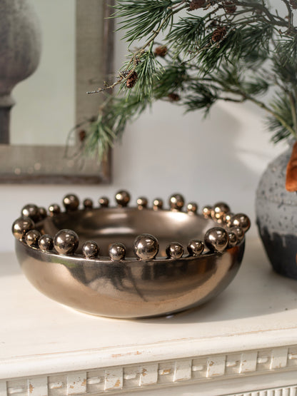 Decorative bronze metallic bowl with spherical embellishments on a surface with greenery in the background