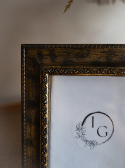 Close-up of an ornate dark brown wooden picture frame with a distressed finish and gold-toned inner trim, holding a white mat with a circular monogram logo.