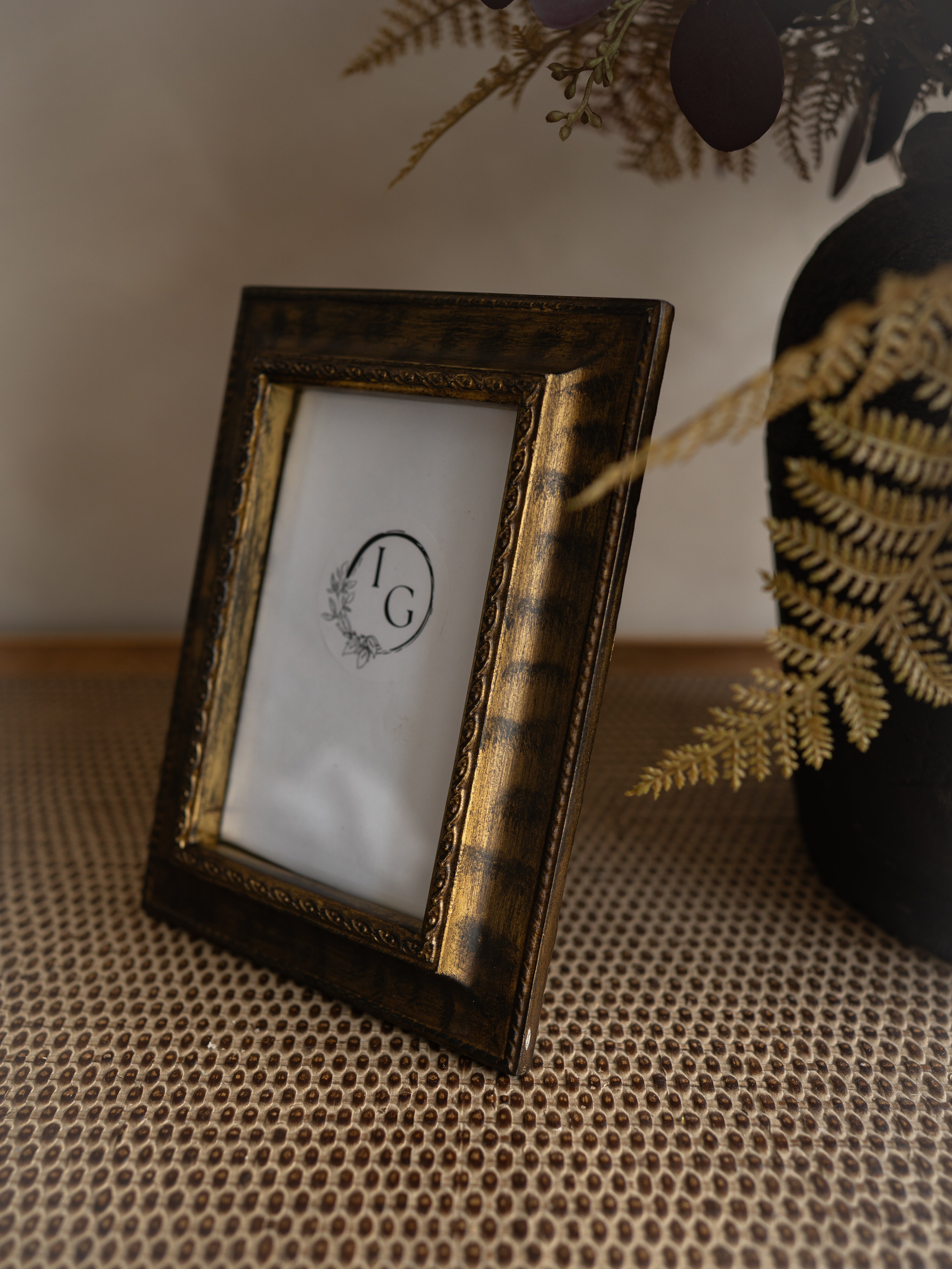 A small rectangular picture frame with an ornate distressed gold and dark brown frame, standing on a woven beige surface. The frame is shown empty with a white insert featuring a circular monogram logo.