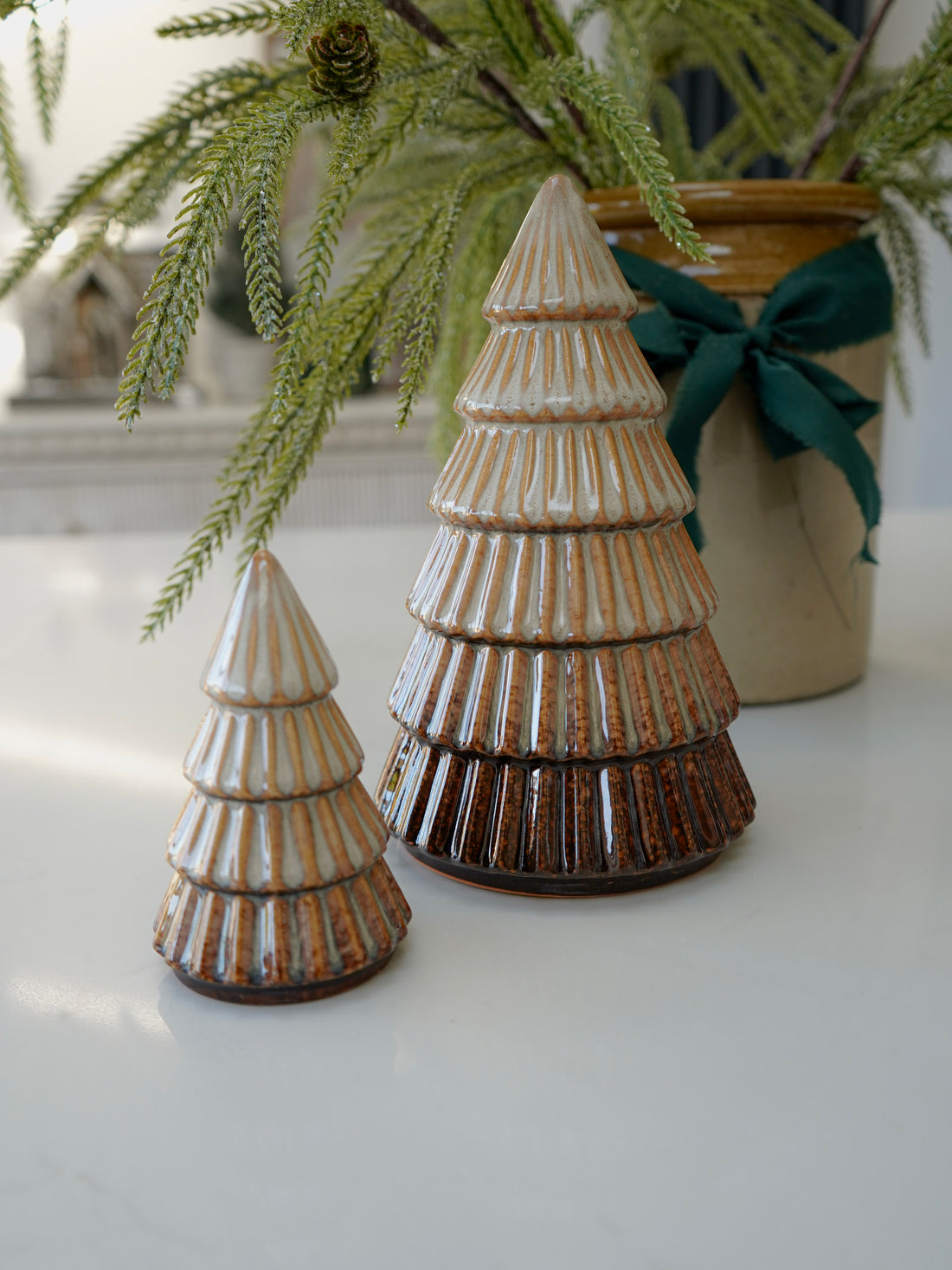 Two ceramic Christmas tree ornaments on a white surface with a plant in the background.