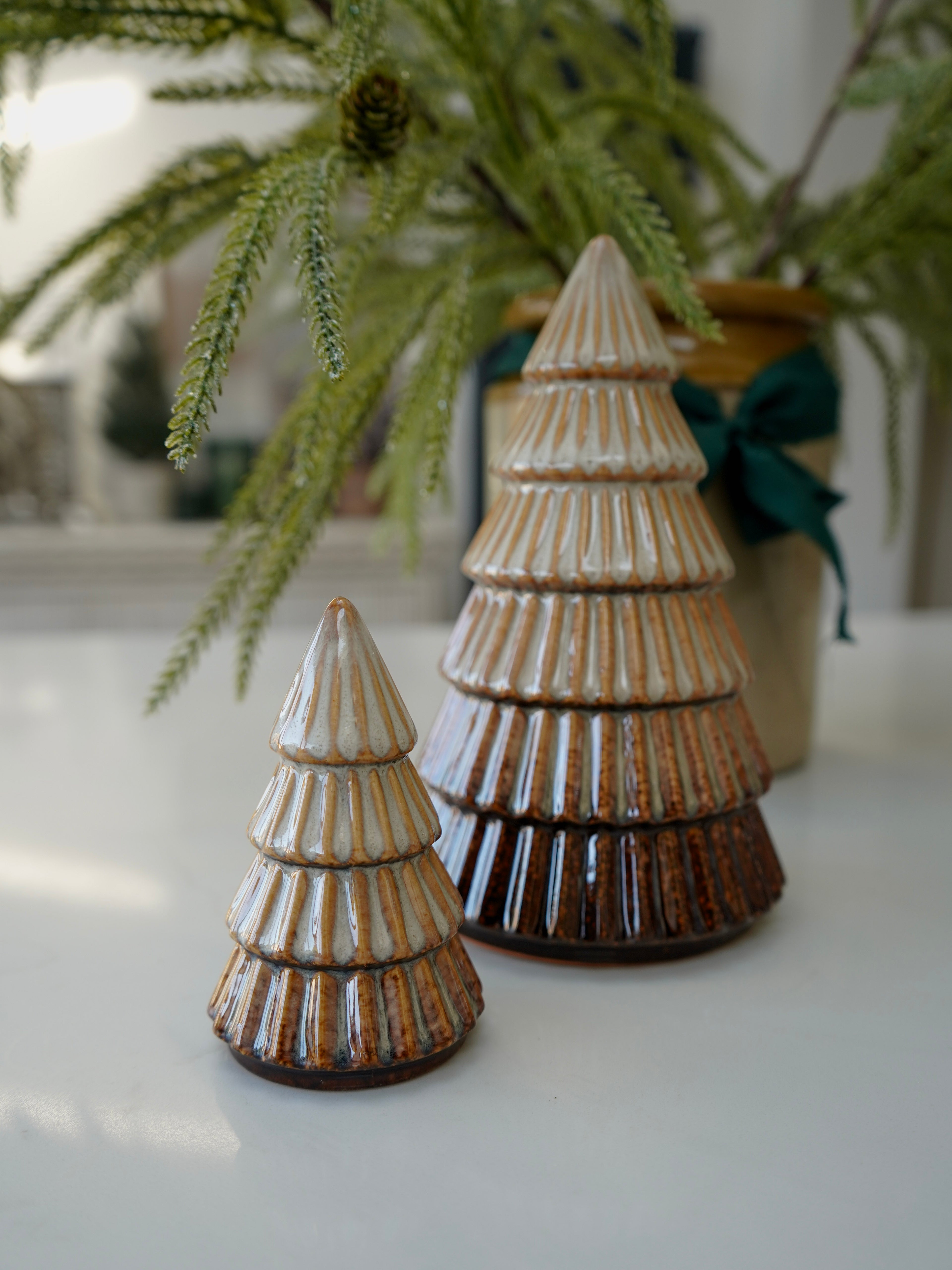 Two ceramic Christmas trees on a white surface with greenery in the background