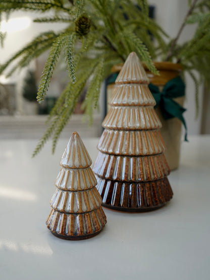 Two ceramic Christmas trees on a white surface with greenery in the background