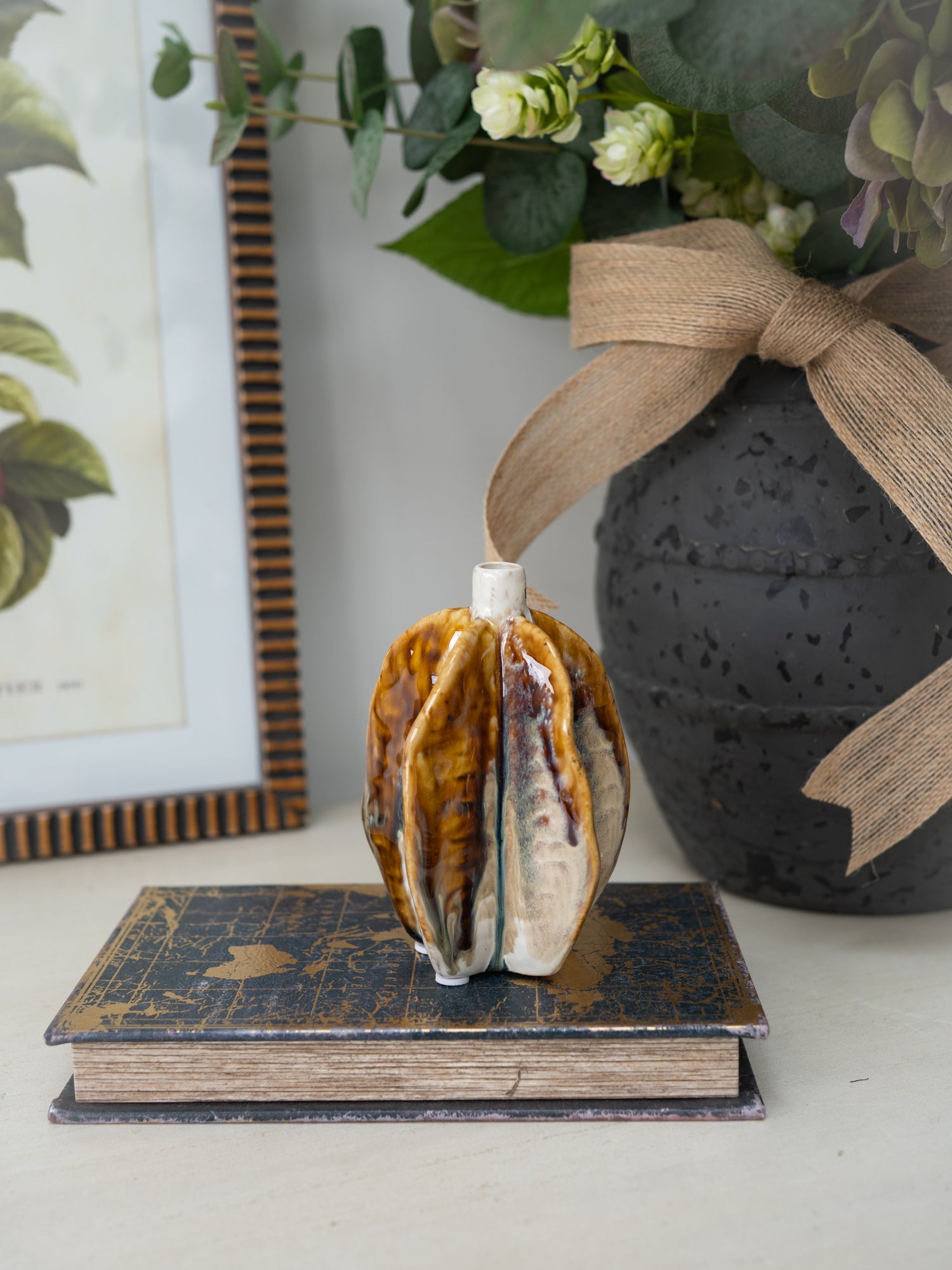 Decorative pod vase on a book with a black vase and greenery in the background