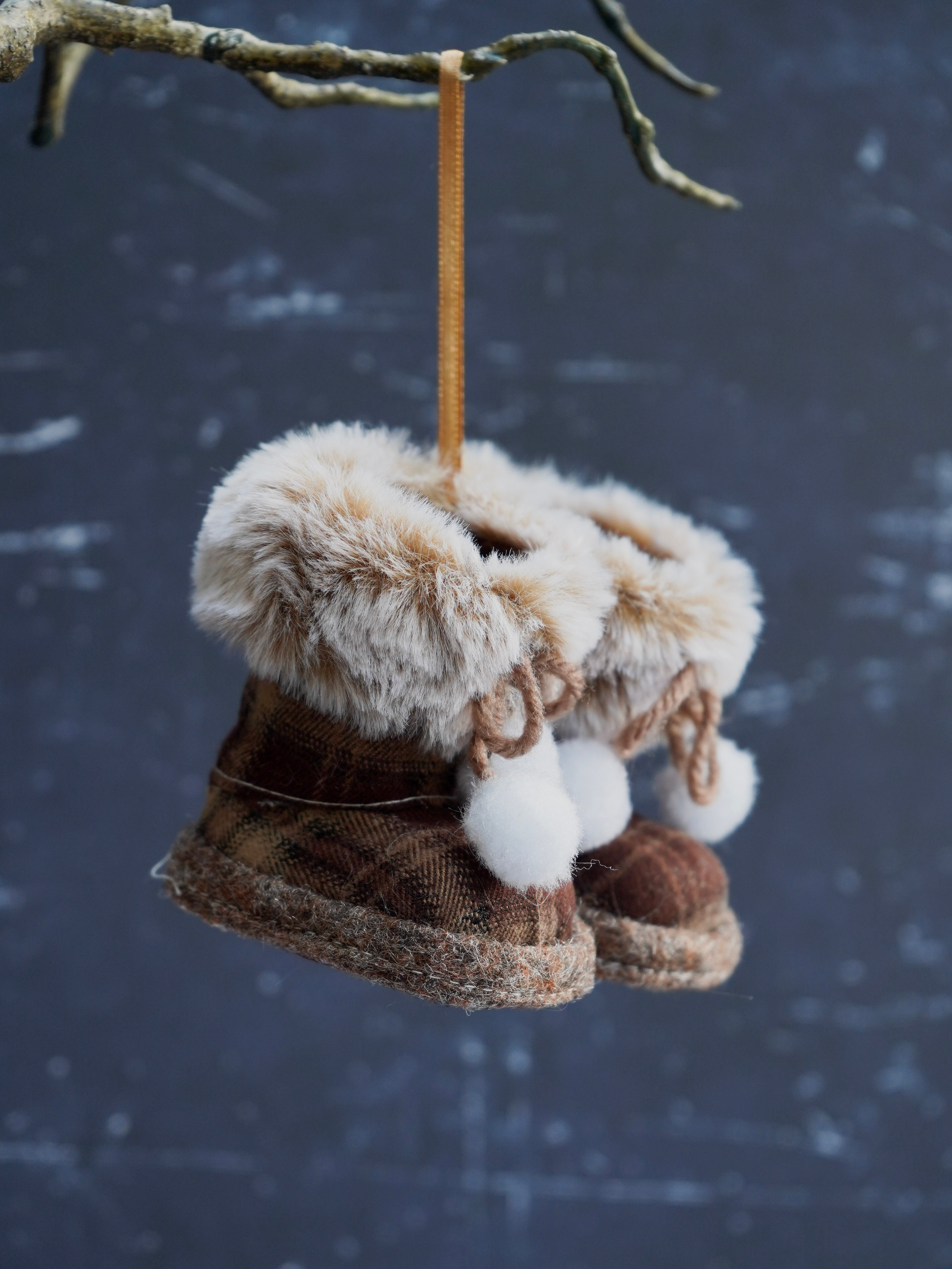 Decorative boots with fur and pom-poms hanging from a branch against a snowy background