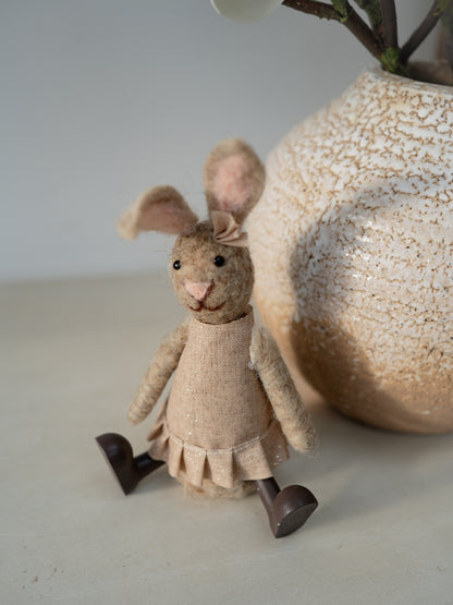 Textile rabbit figure in a dress next to a textured vase on a light surface.