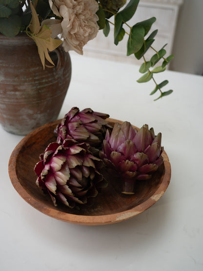 Three faux whole artichokes with deep purple and green tones arranged in a small round wooden bowl on a white tabletop, with a rustic brown vase and dried flowers blurred in the background.