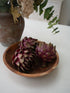 Three faux whole artichokes with deep purple and green tones arranged in a small round wooden bowl on a white tabletop, with a rustic brown vase and dried flowers blurred in the background.