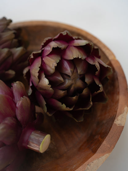 A close-up of a  faux dark purple artichoke inside a rustic wooden bowl, with other artichokes partially visible on the left against a light background.