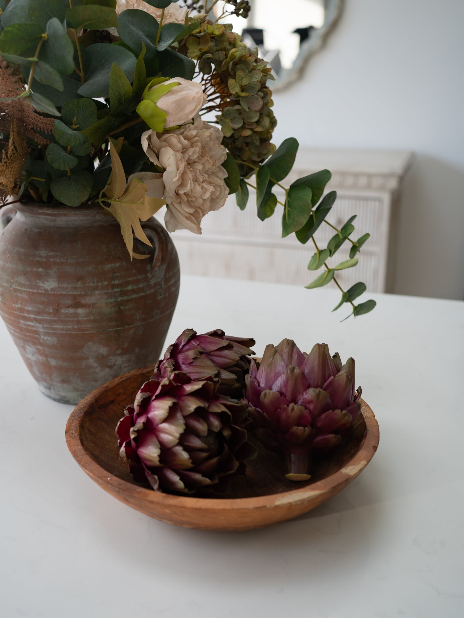 A rustic wooden bowl containing three faux purple artichokes on a white table, with a large terracotta vase of flowers in the background.