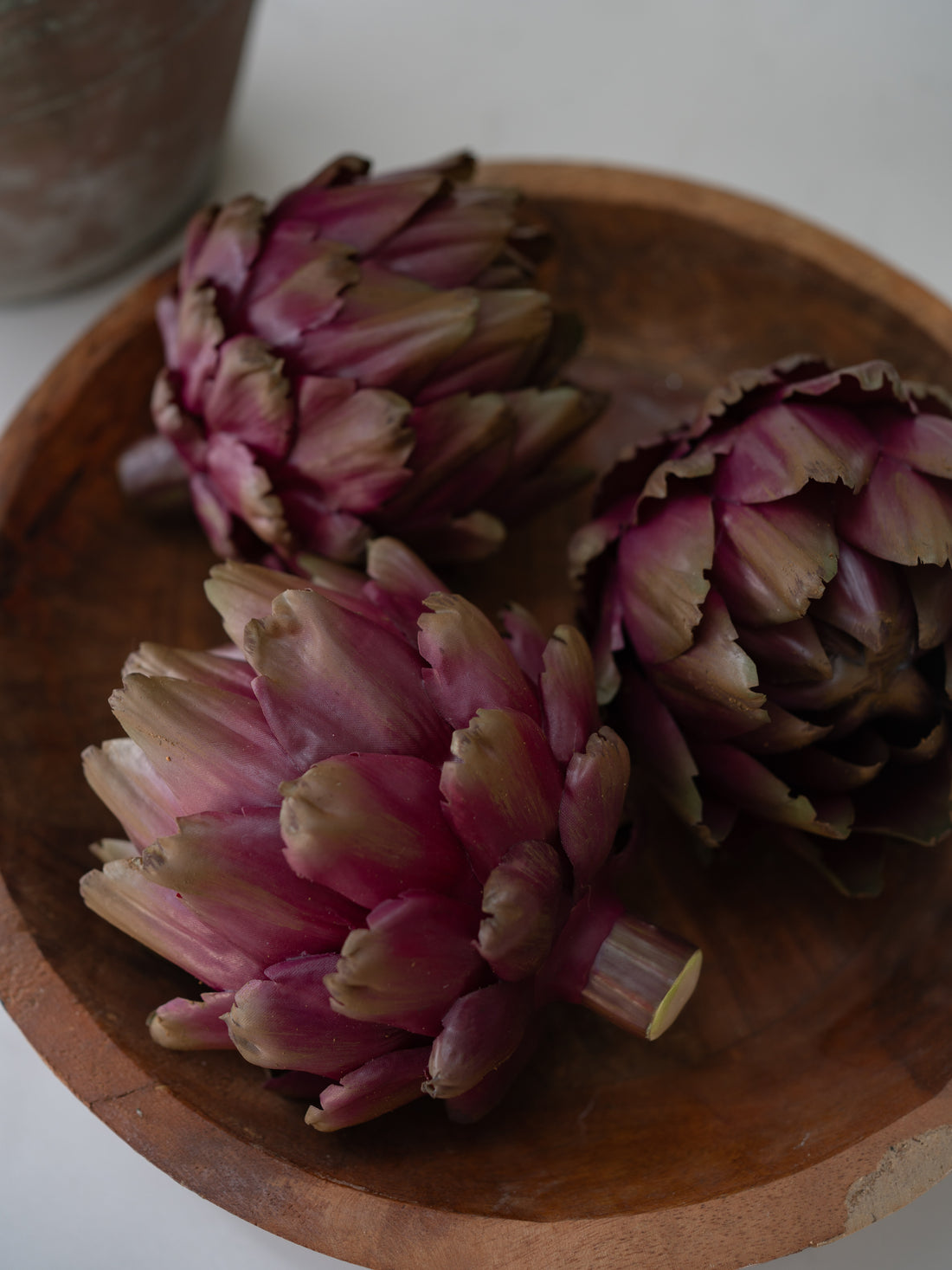 Three whole  faux artichokes with pinkish-purple and pale beige tones arranged on a round wooden plate.