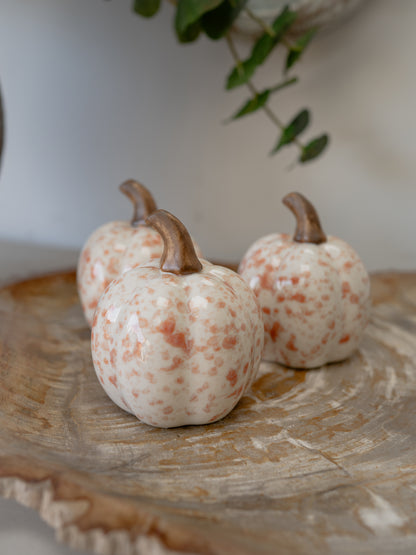 Three small decorative ceramic pumpkins with speckled orange and cream glazes and brown stems, arranged on a rustic wood slice tray with a soft-focus indoor background.