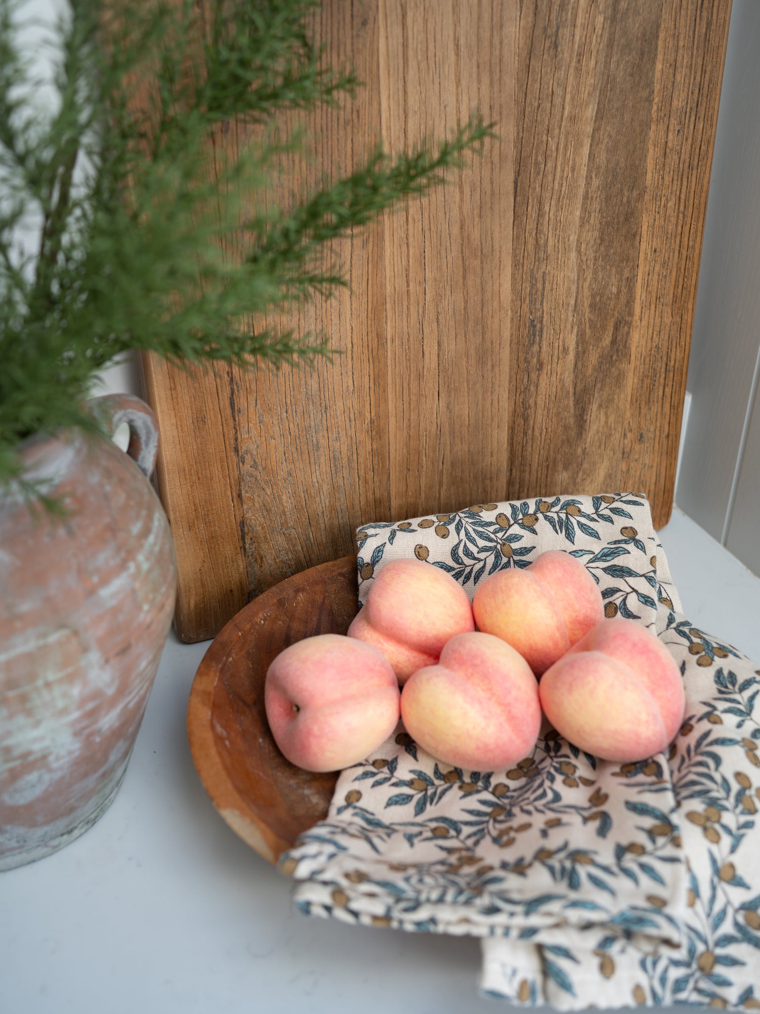 Bowl of faux decorative peaches on a floral cloth with a wooden cutting board and plant in the background