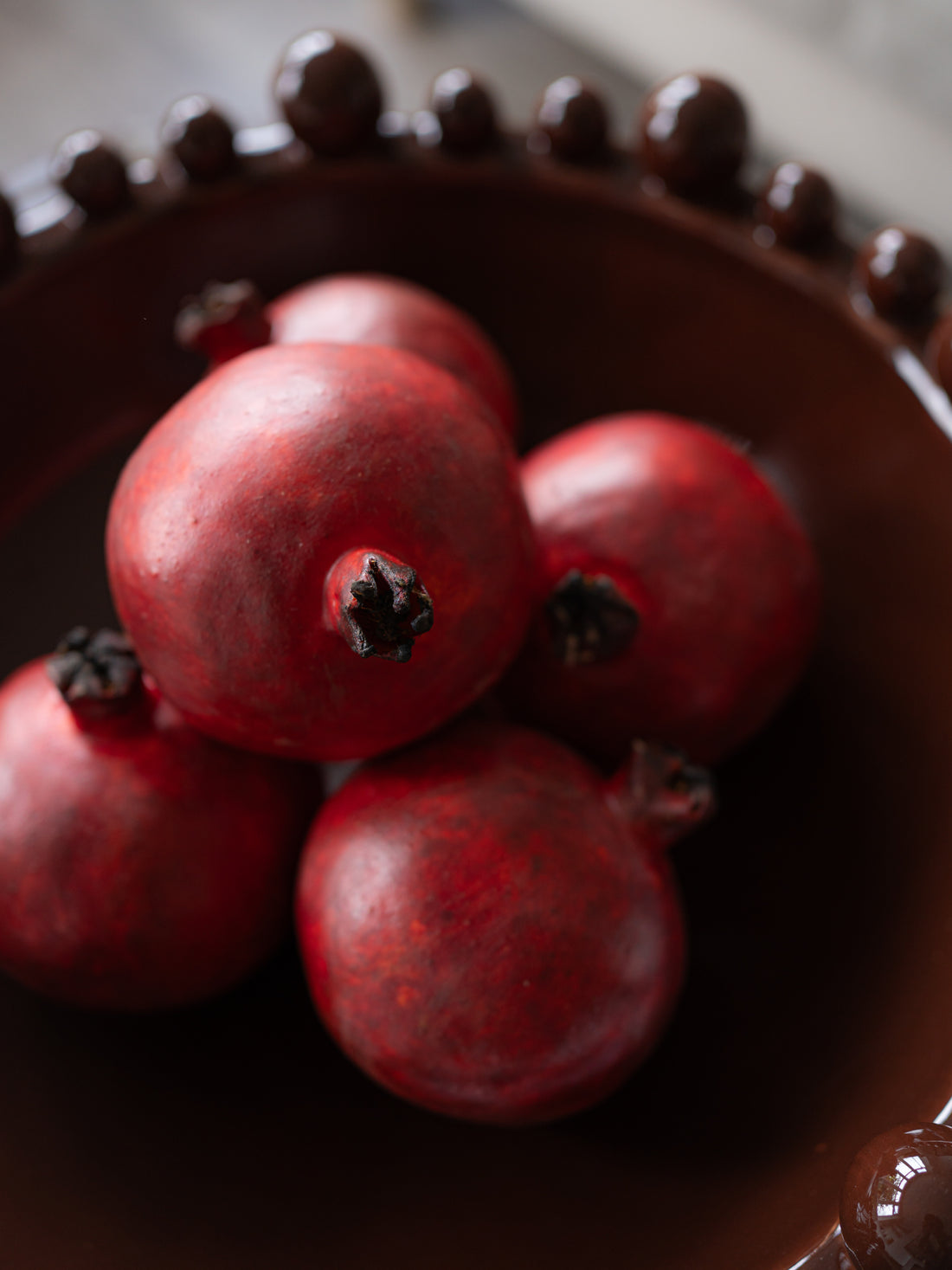 Several glossy red pomegranates resting in a dark brown bowl with a beaded rim, photographed in a close-up still life.