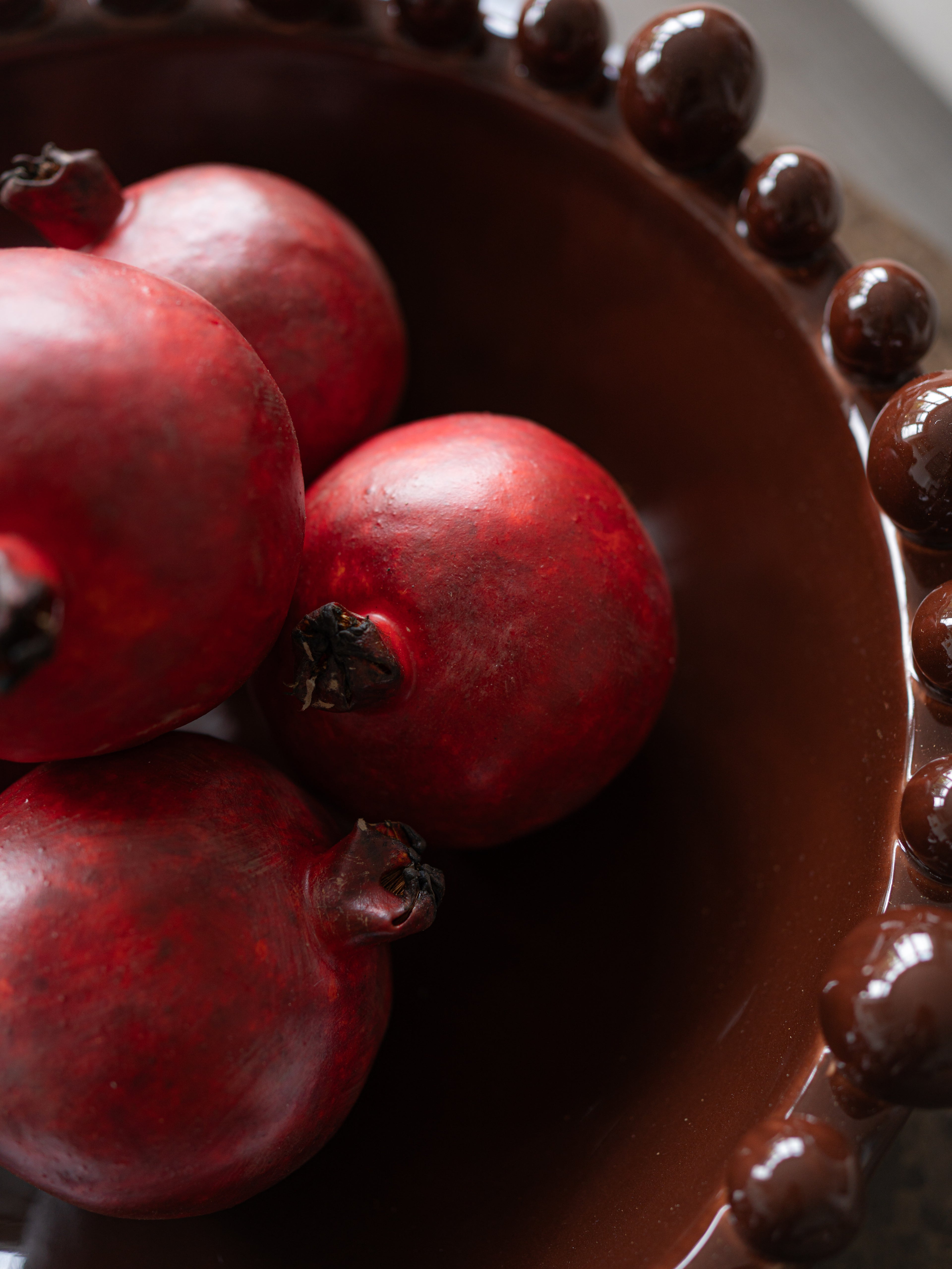 Several glossy faux red pomegranates resting inside a dark brown beaded bowl.