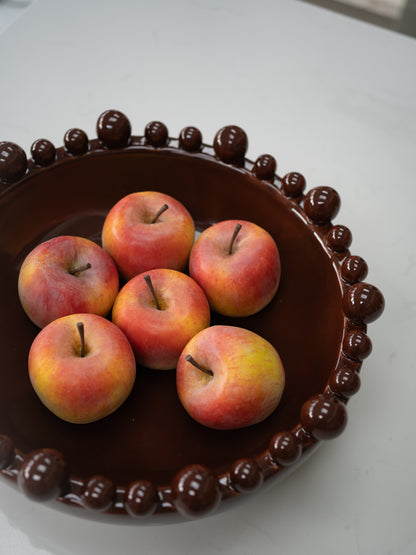 Six faux red and yellow apples arranged inside a dark brown ceramic bowl with a beaded rim, set on a light gray surface.