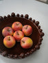 Six faux red and yellow apples arranged inside a dark brown ceramic bowl with a beaded rim, set on a light gray surface.