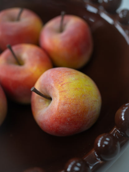 A close-up shot of several faux red and yellow apples resting in a dark brown ceramic bowl with a beaded rim.