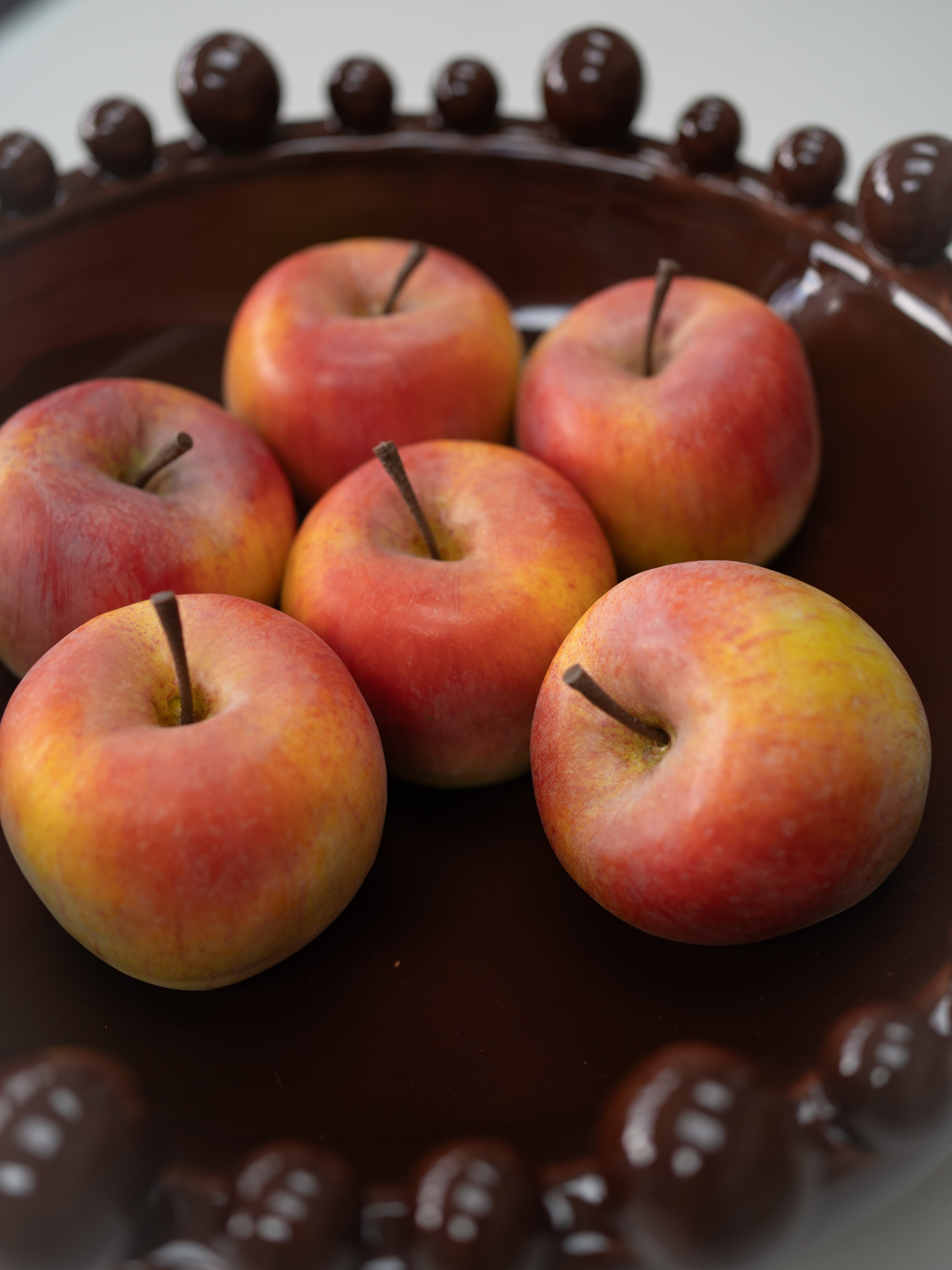 Six faux red and yellow apples arranged on a dark brown ceramic plate with a decorative beaded rim.