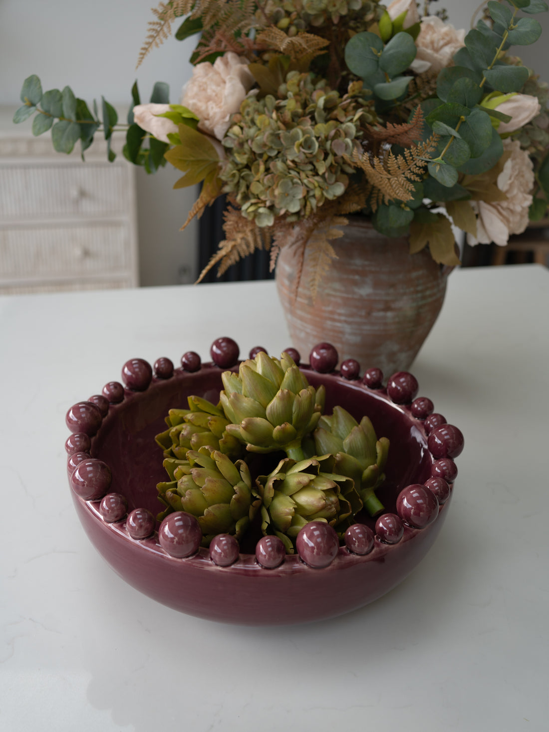 A round burgundy ceramic bowl with a raised beaded rim, filled with green artichokes, set on a white tabletop with a floral arrangement in the background.