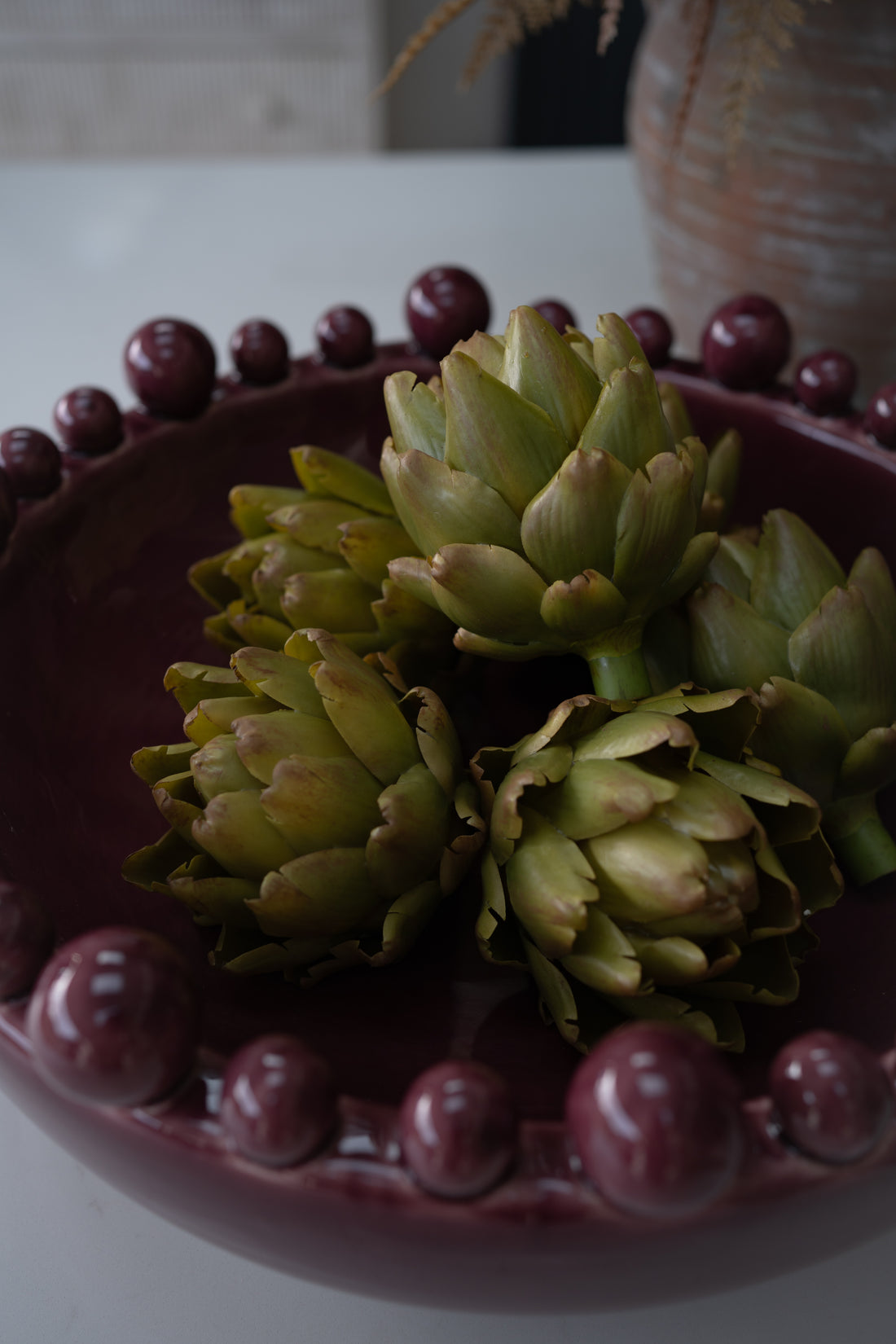Faux green artichokes arranged in a dark burgundy ceramic bowl with a beaded rim, set on a light tabletop with a softly blurred background.
