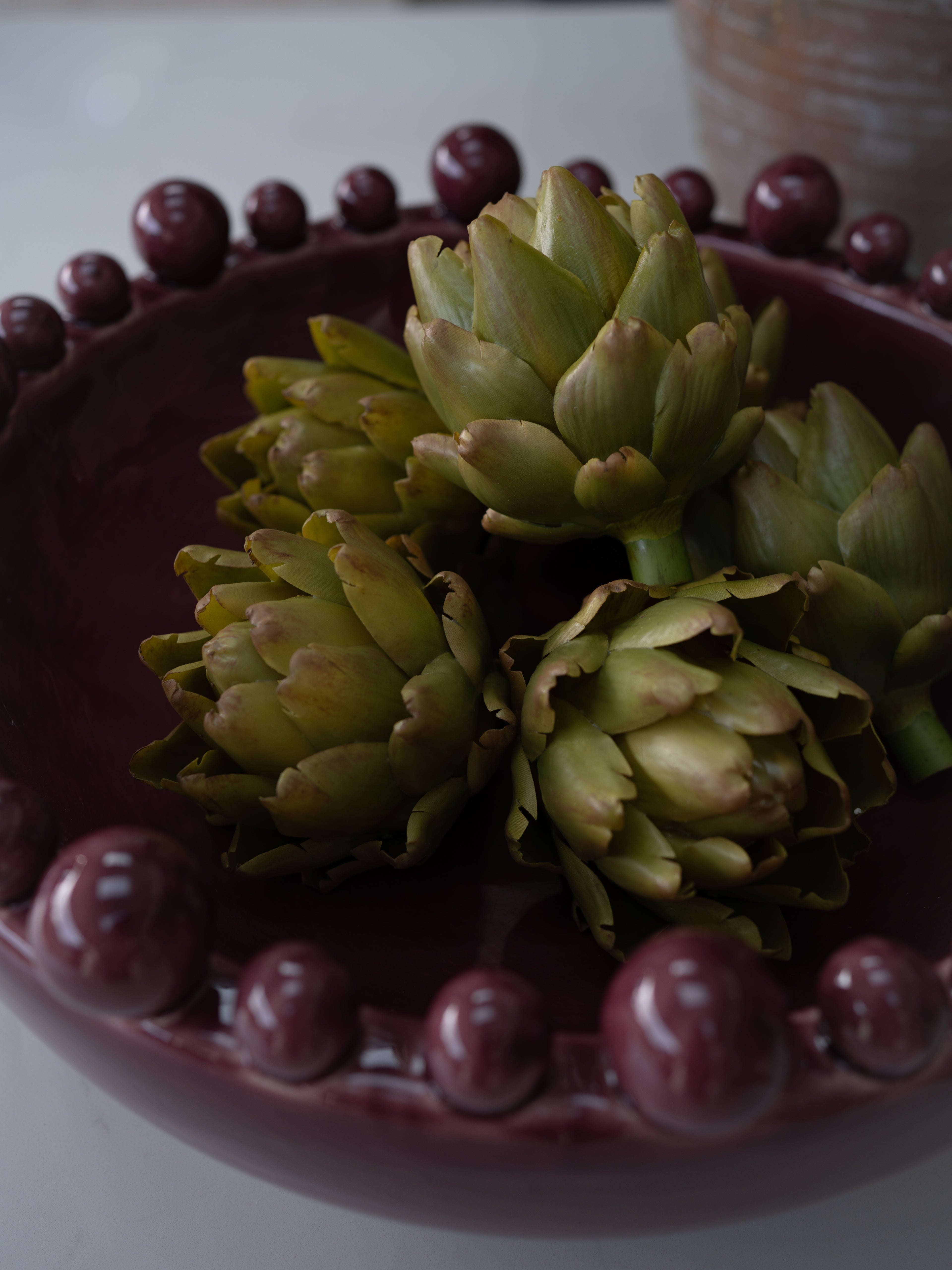 Three faux green artichokes placed in a dark maroon ceramic bowl with a beaded rim, photographed on a light surface with a softly blurred background.