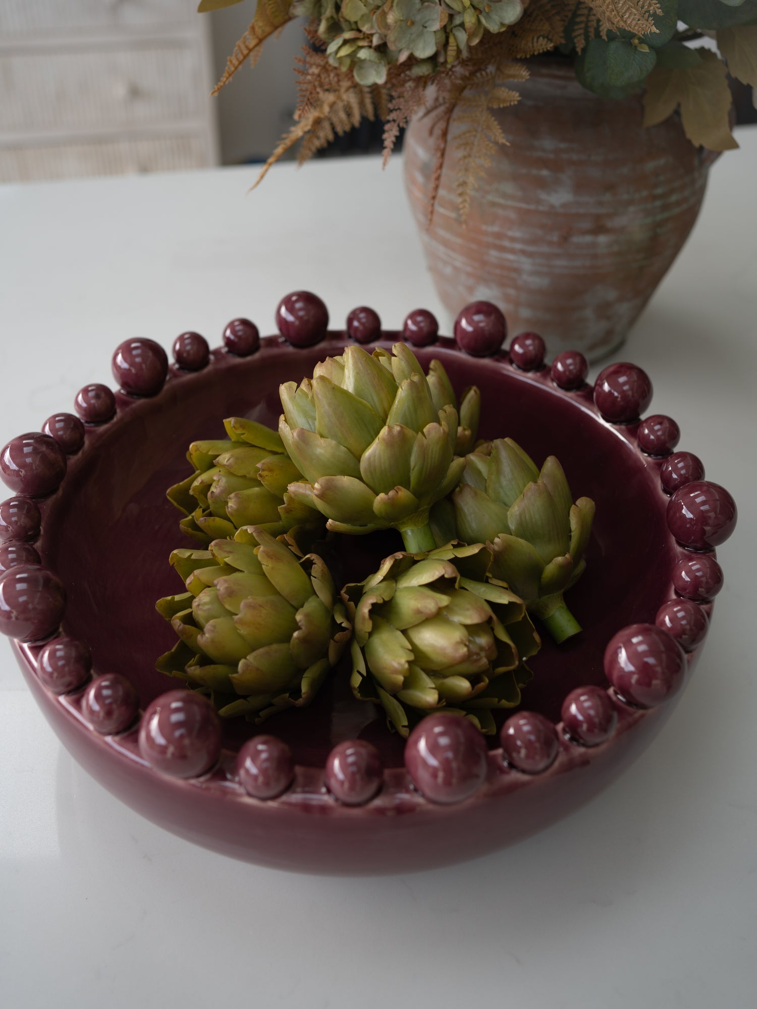 A round maroon ceramic bowl with a beaded rim, containing three faux green artichokes, set on a white surface with a blurred rustic vase in the background.