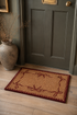 A rectangular coir doormat with a dark red border and bow pattern, placed on a light wood floor in front of a dark green door.