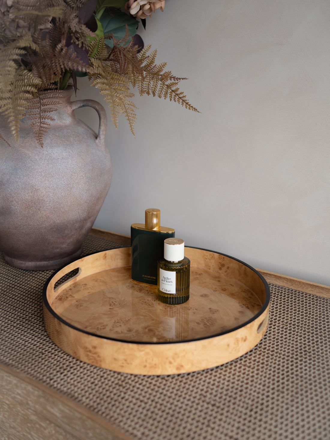 Burl wood effect tray with perfume bottles on a textured surface, with a vase and plants in the background.