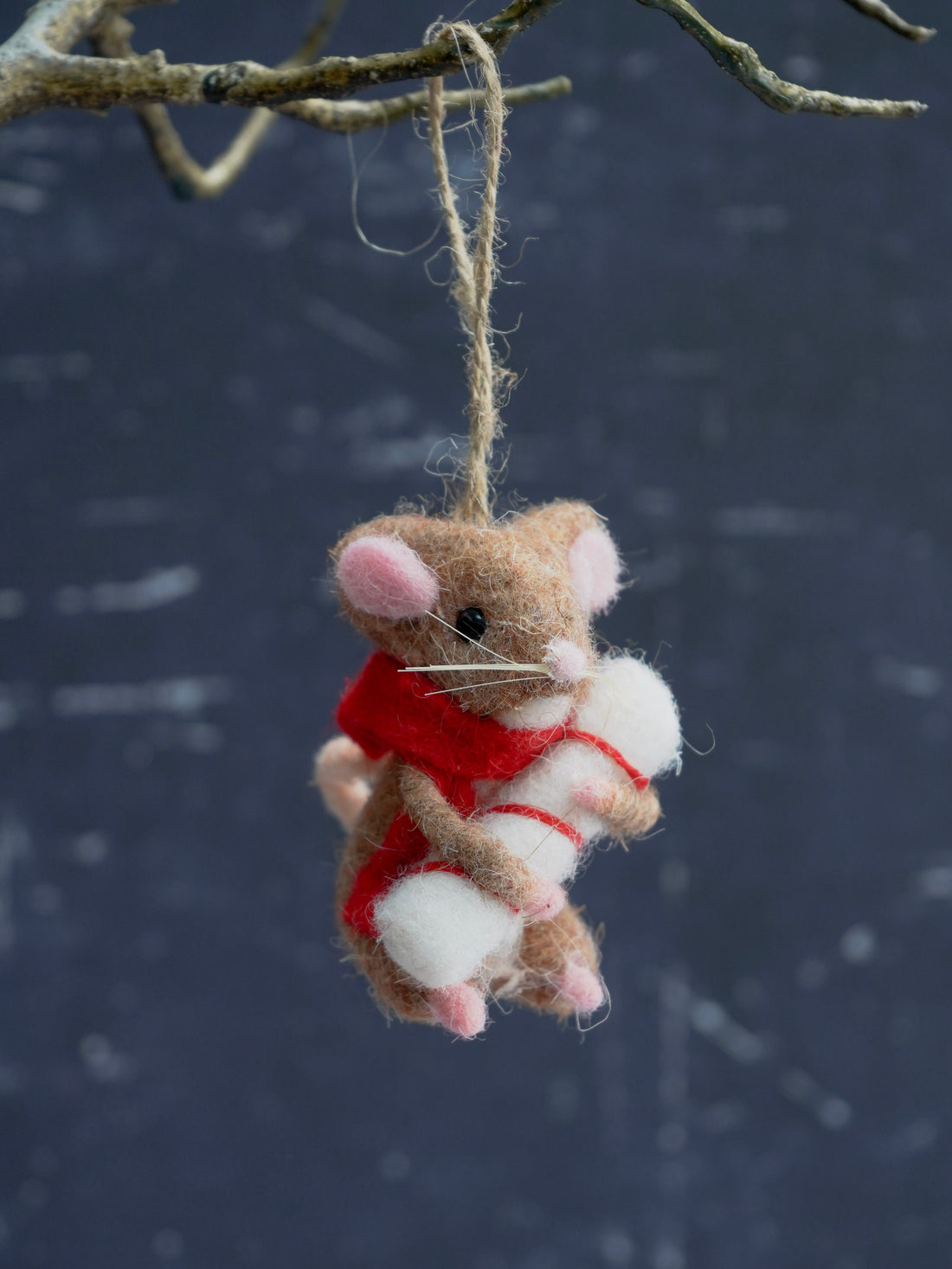 Felted mouse ornament with a red scarf hanging from a branch against a dark background.