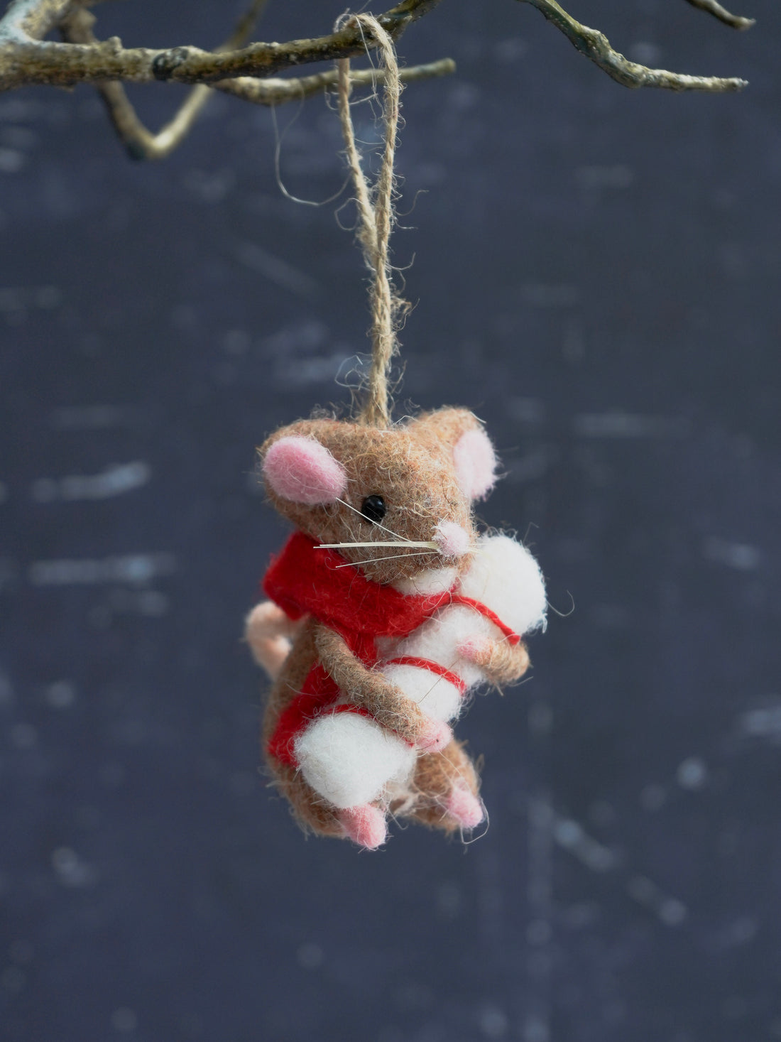 Felted mouse ornament with a red scarf hanging from a branch against a dark background.