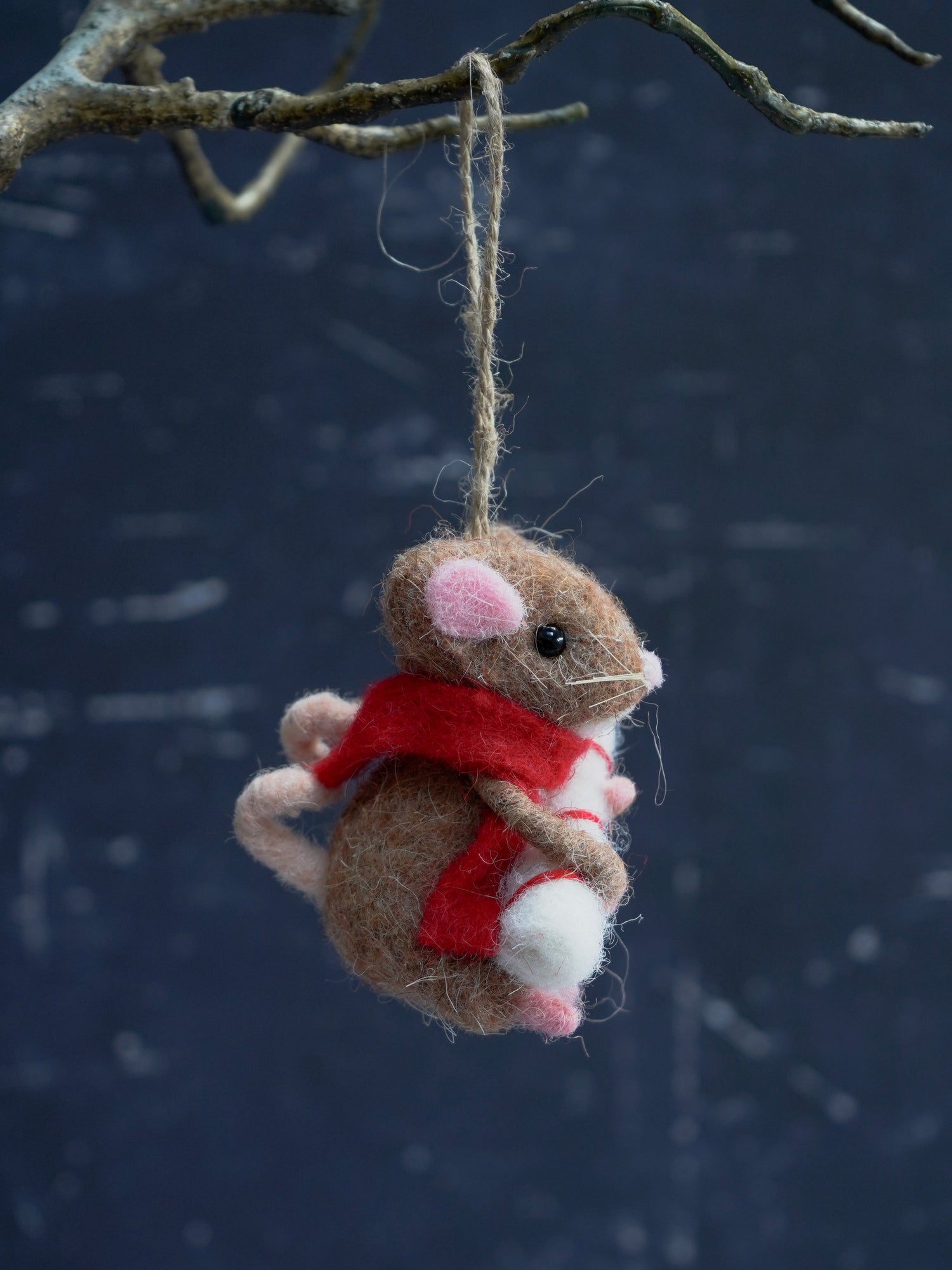 Felted mouse ornament with a red scarf hanging from a branch against a dark background.