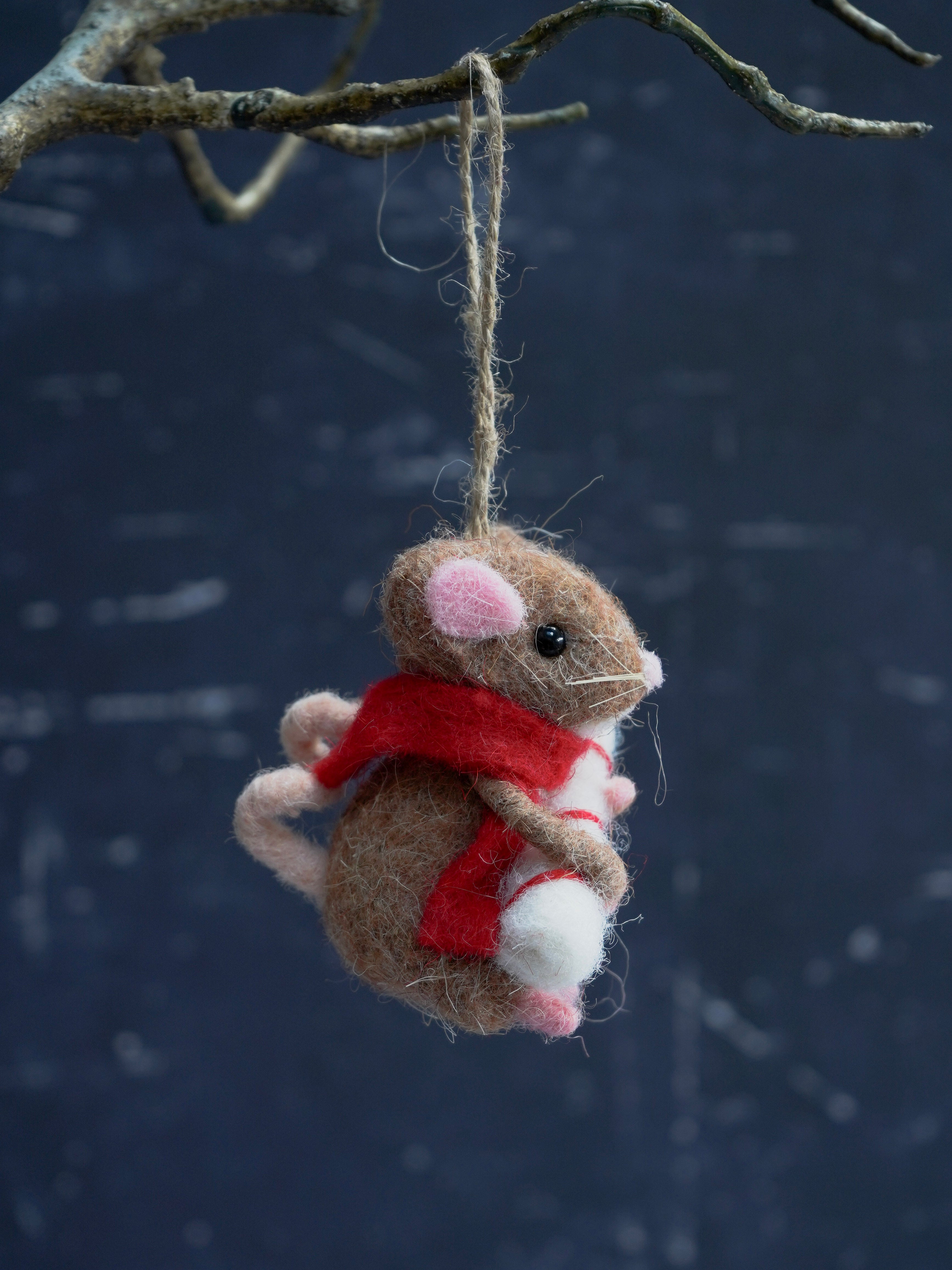 Felted mouse ornament with a red scarf hanging from a branch against a dark background.