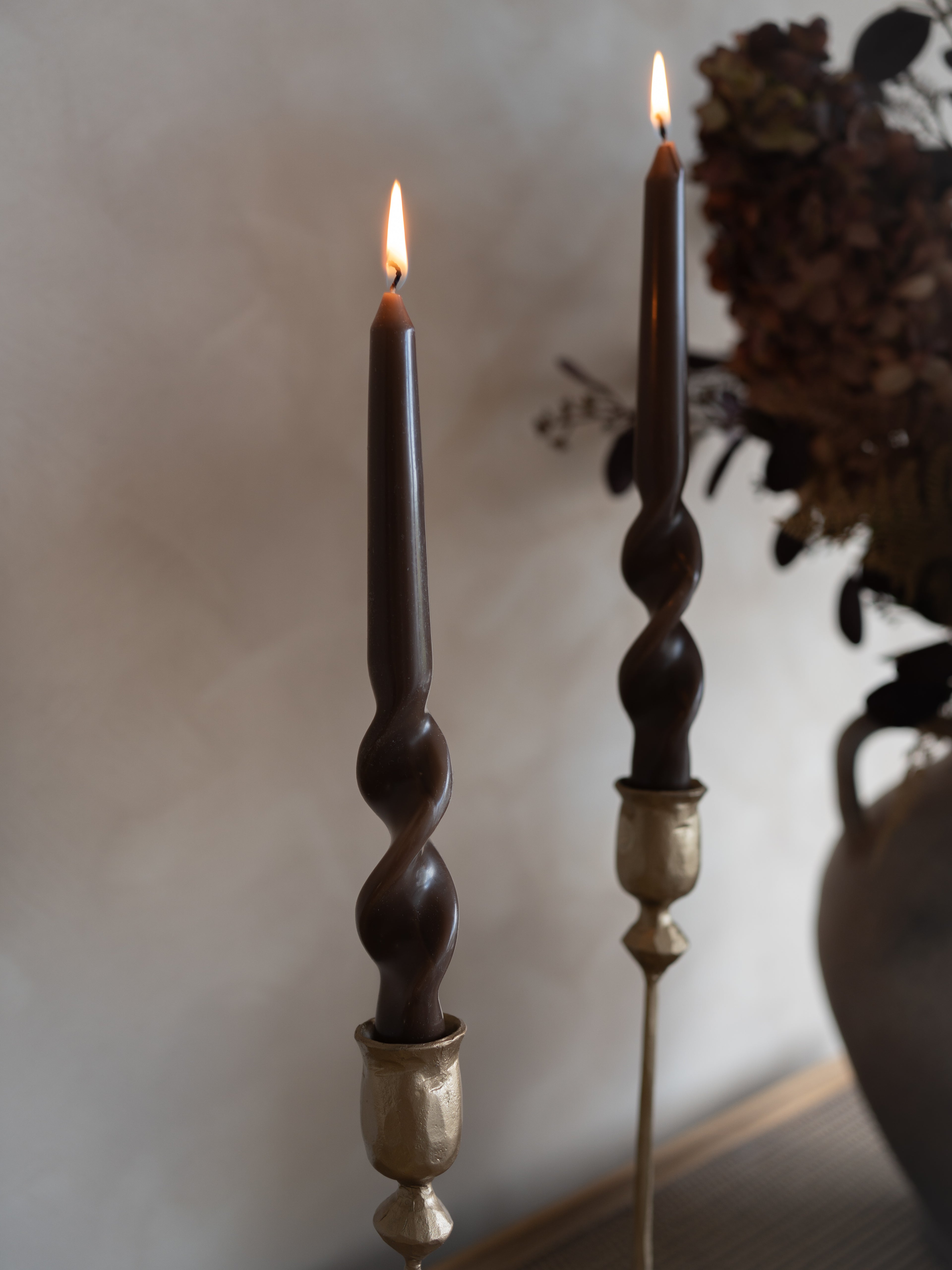 Two dark caramel dinner candles with twisted designs and gold bases on a neutral background.
