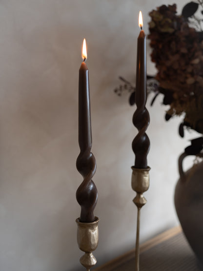 Two dark caramel dinner candles with twisted designs and gold bases on a neutral background.