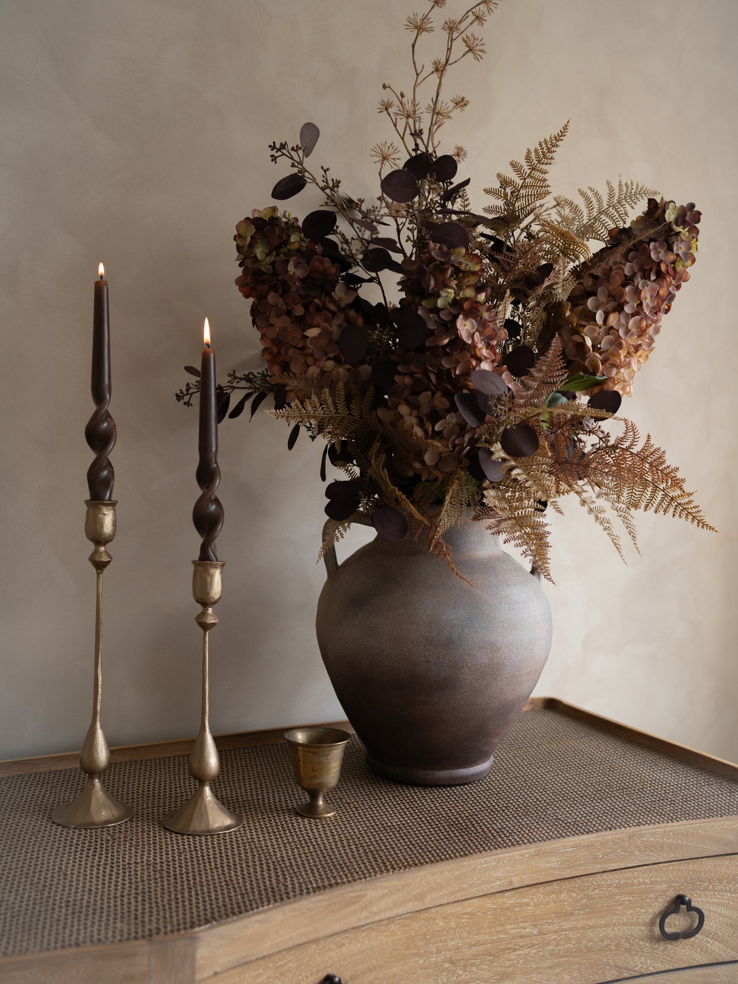 Decorative arrangement with a vase of dried flowers, candles, and a small cup on a textured surface.