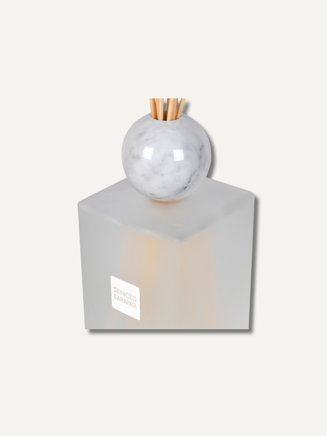 A reed diffuser bottle with a frosted translucent square base, a round white marble sphere top, and natural reed sticks inserted at the top, shown on a light beige background. A small white label on the front reads &