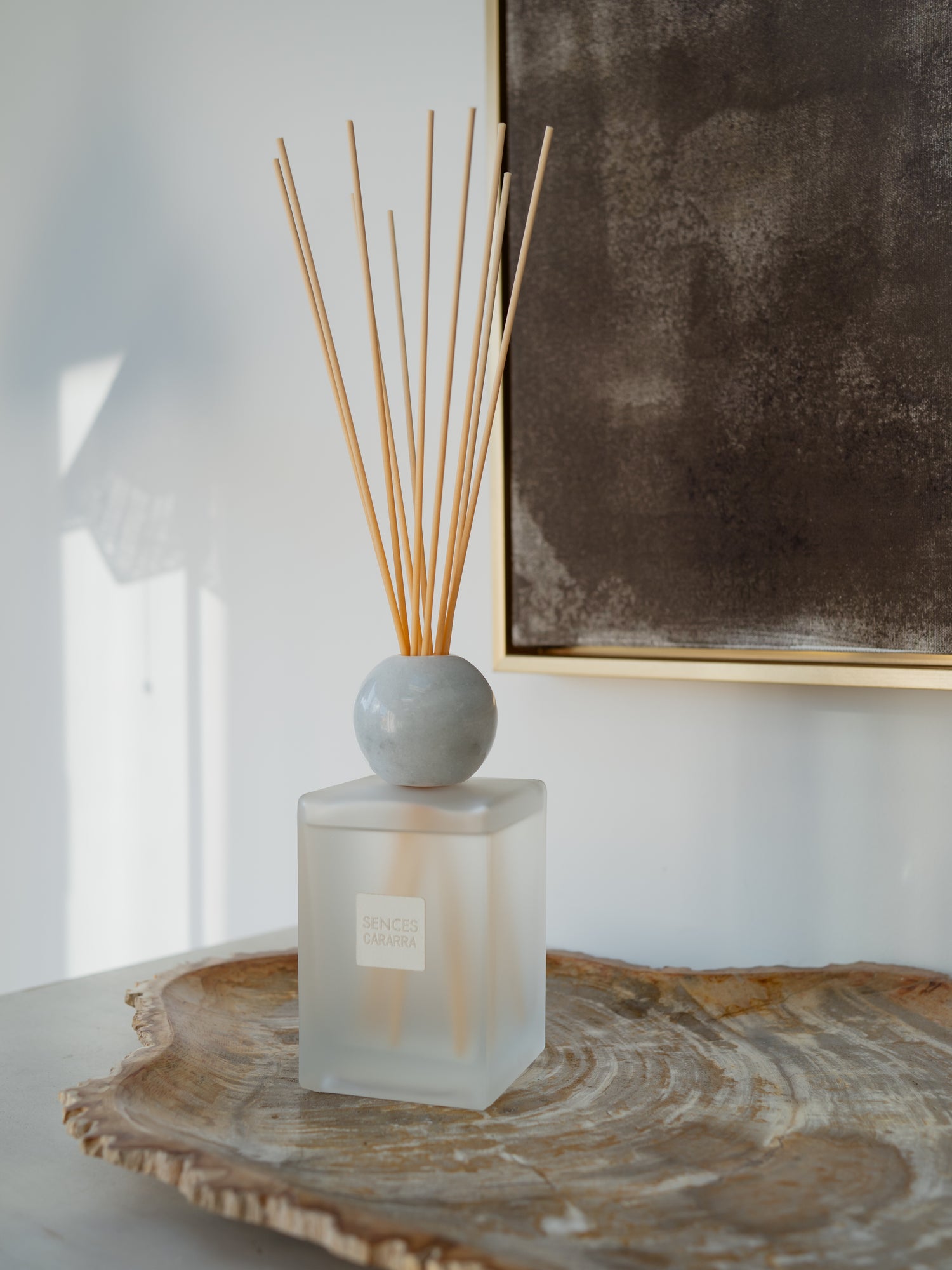 A reed diffuser bottle with a frosted glass base, a round white marble stopper, and natural reed sticks, styled on a rustic wood slice tray against a white wall with a framed artwork.