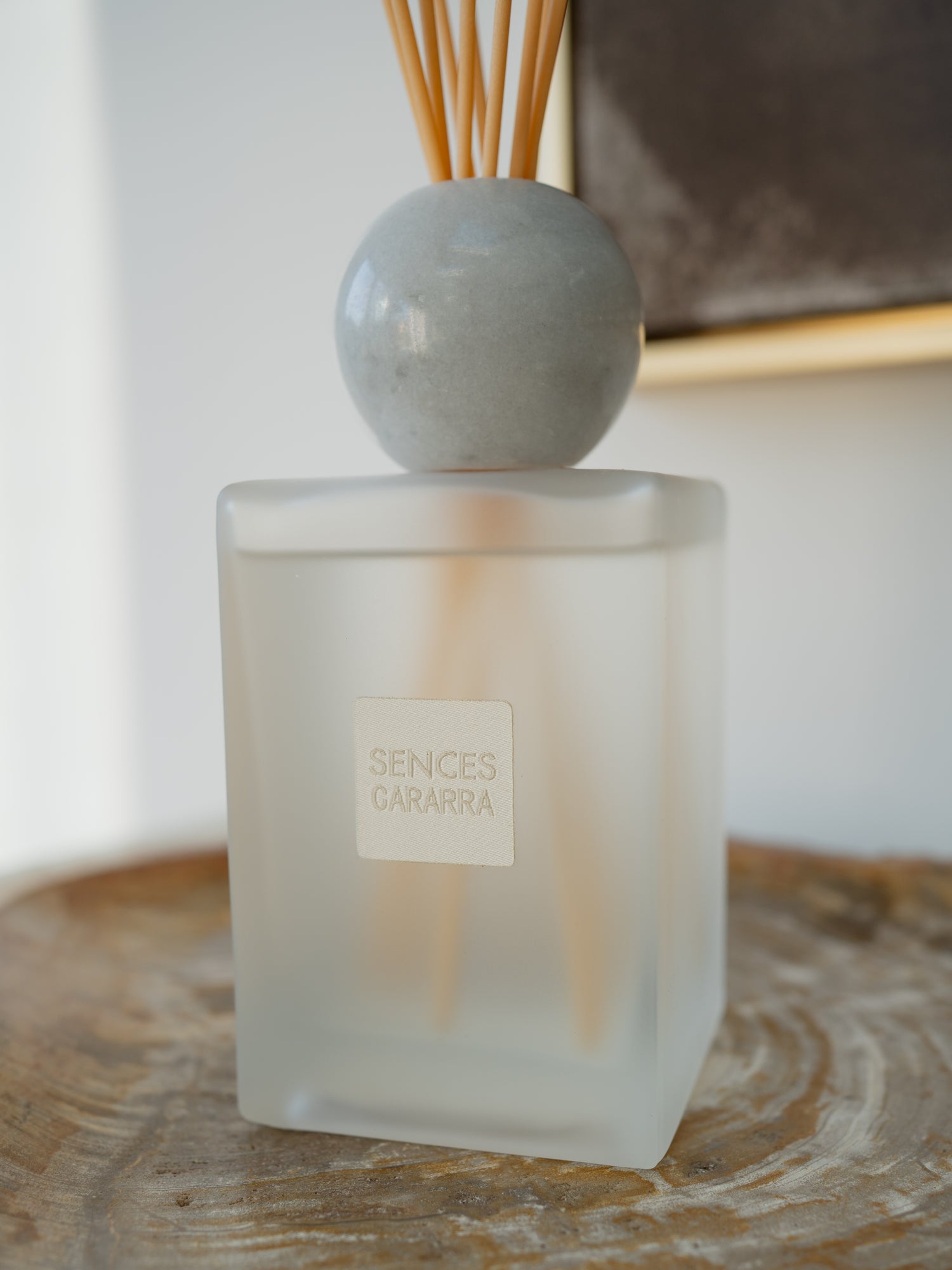 A frosted glass reed diffuser bottle with a round gray marble stopper and natural wood reeds, sitting on a rustic wooden slice. The front label reads &