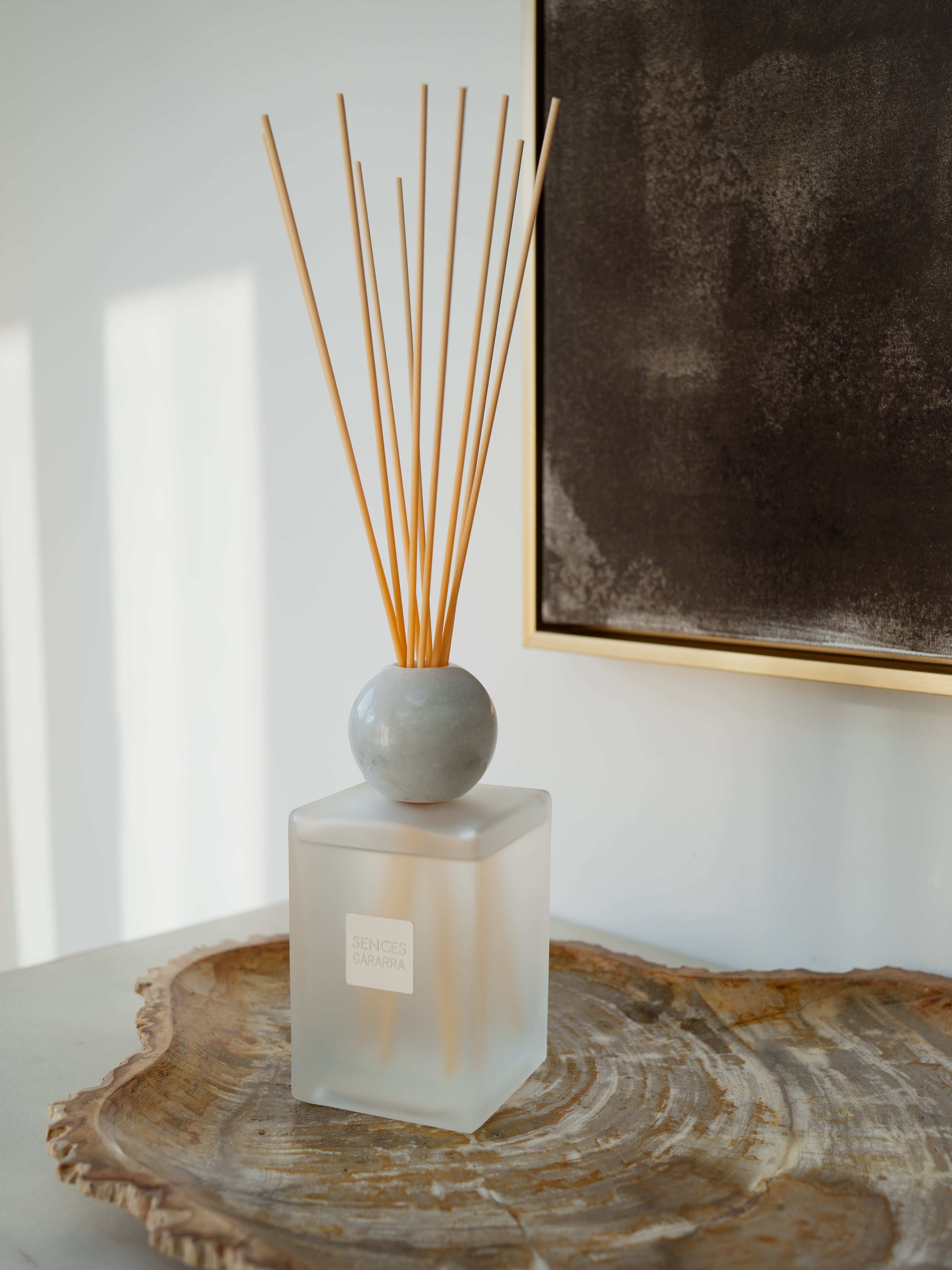 A reed diffuser with a frosted square glass bottle, a round gray ceramic stopper, and natural wood reeds, displayed on a rustic wood slice tray against a white wall.