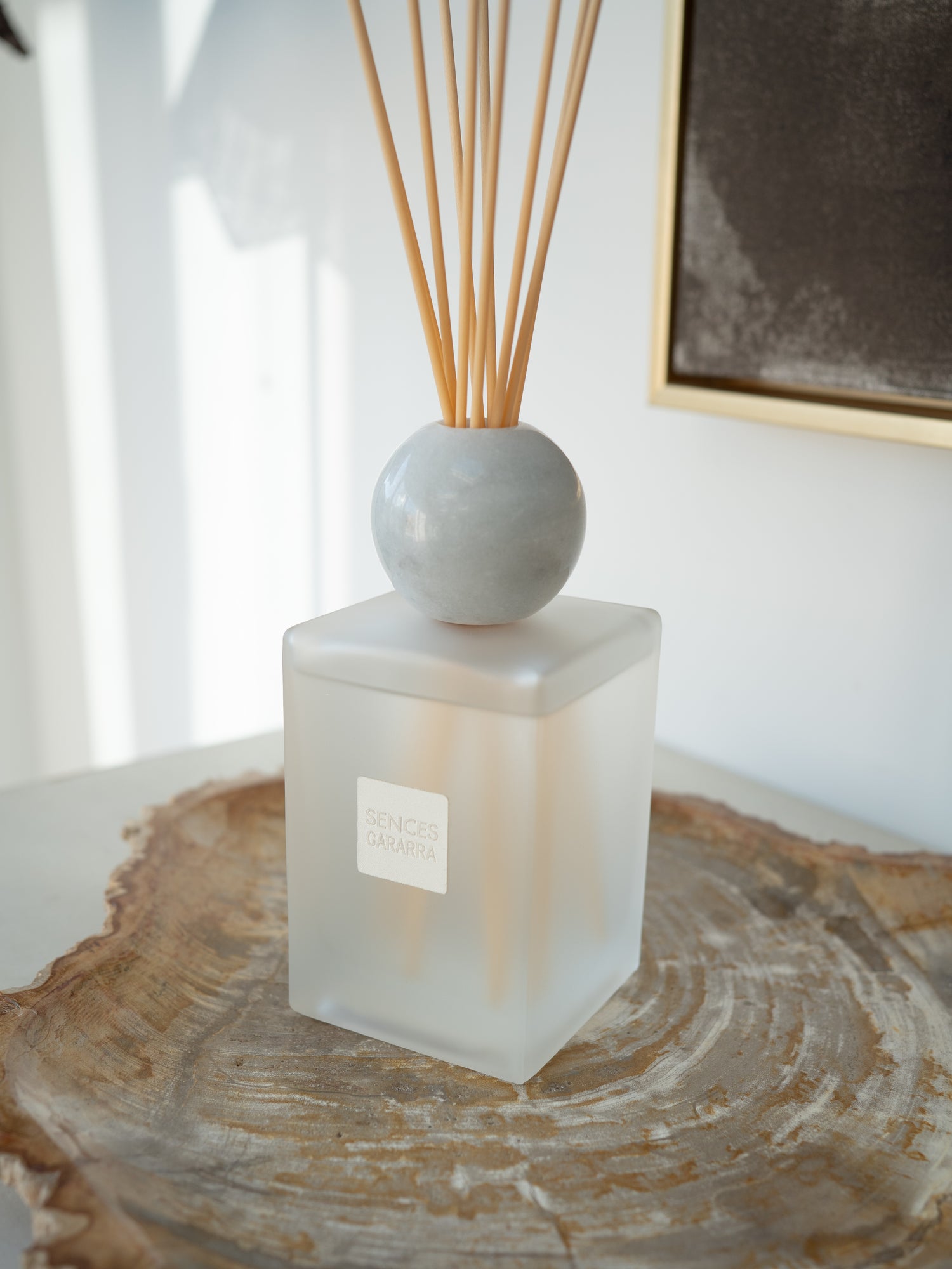 A reed diffuser with a frosted square glass bottle, a round light WHITE marble stopper, and natural wood reeds, displayed on a rustic wood slice slice in a bright, modern room.