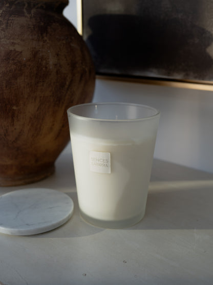 A frosted glass candle jar filled with white wax, shown next to its white marble lid on a light surface. A small label on the jar reads &