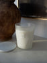 A frosted glass candle jar filled with white wax, shown next to its white marble lid on a light surface. A small label on the jar reads &