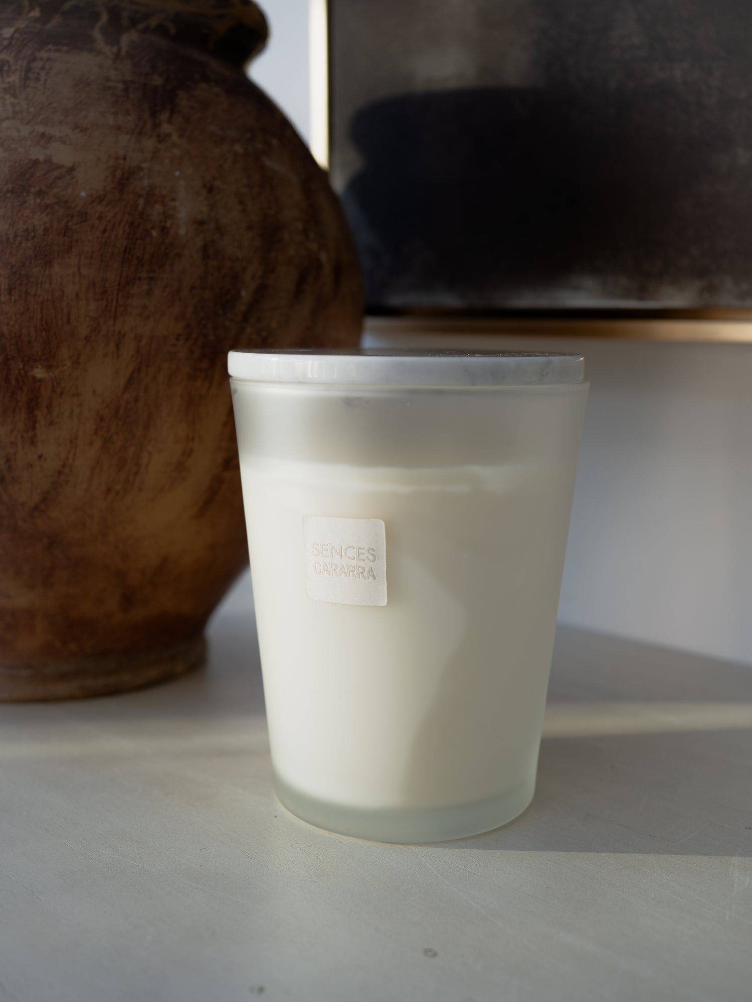 A frosted glass jar candle with white wax sits on a light surface next to a large brown ceramic vase. The candle has a small white label on the front with the brand name &